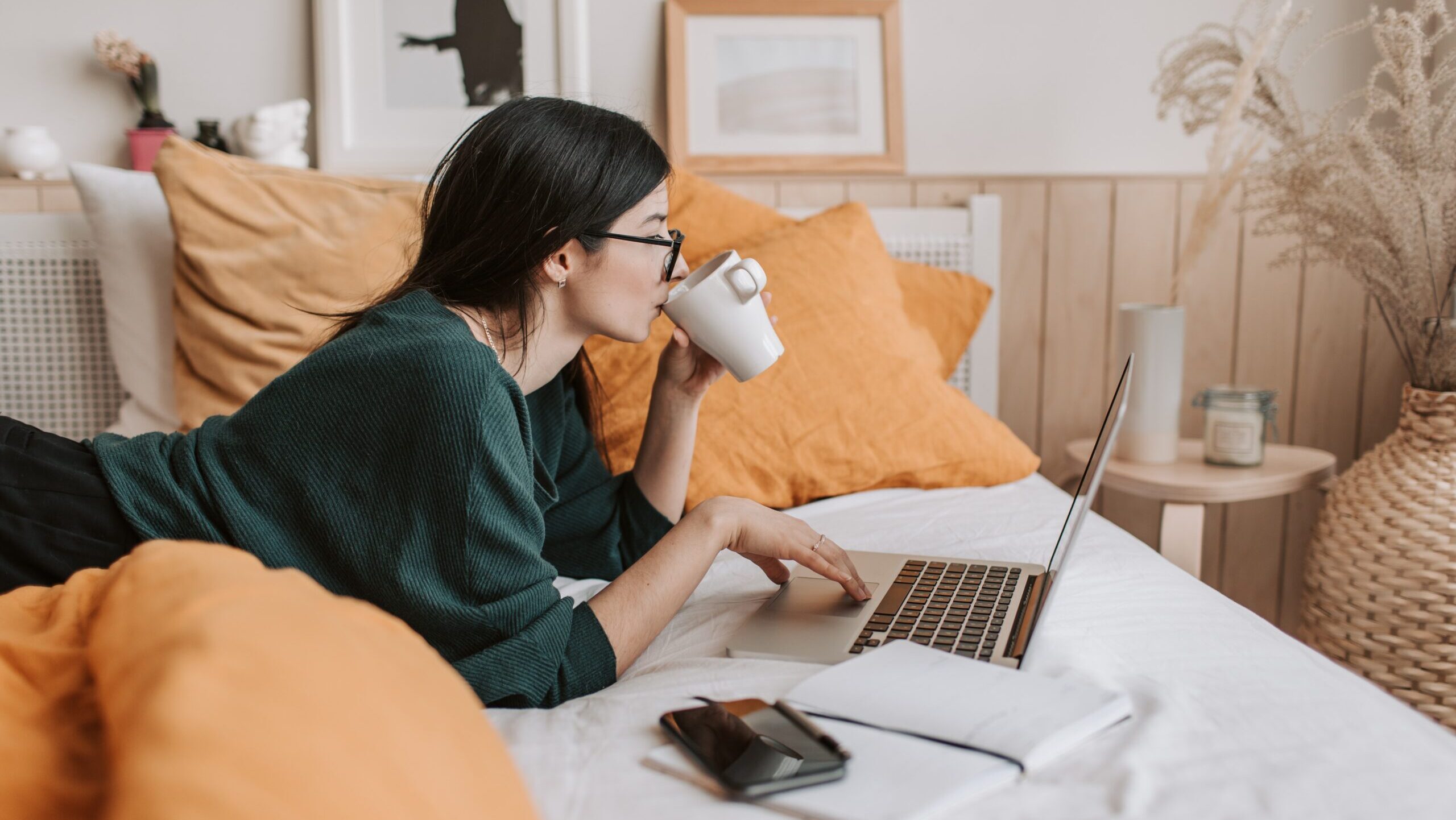 A woman laid back on a bed with a laptop and a cup of coffee.