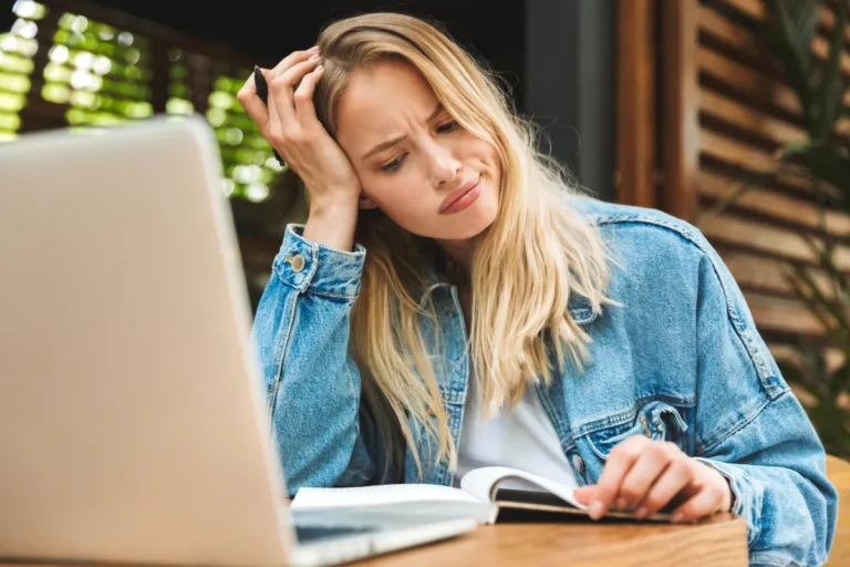 Woman appears puzzled or concerned while looking at a laptop screen and holding a notebook.