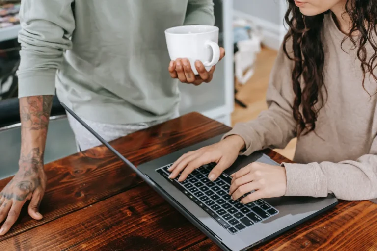 Two individuals collaborating at a wooden table, one typing on a laptop, the other holding a white mug.