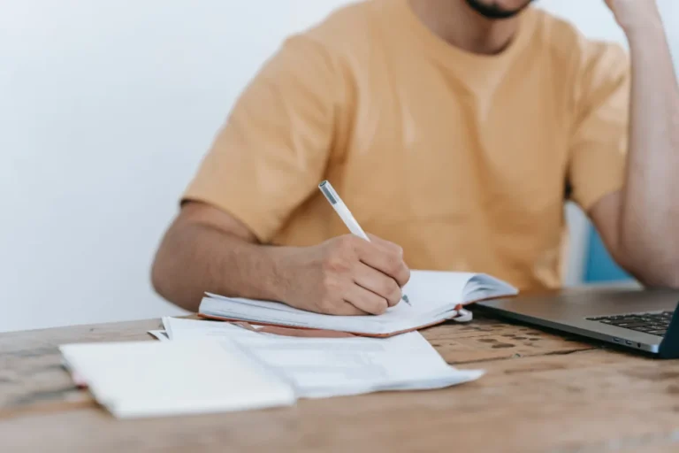 A person sitting at a table writing in a notebook with a laptop and papers nearby.