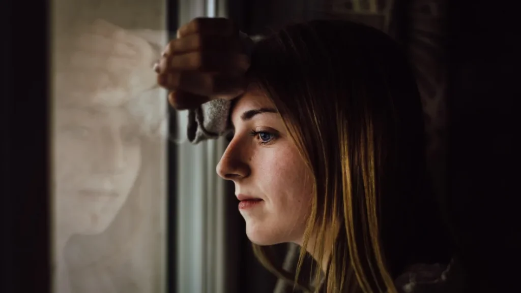 A person gazing outside through a window with their reflection visible on the glass.