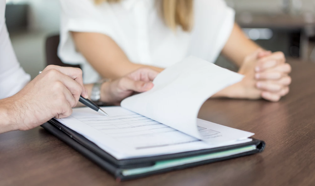 Two people are seated at a desk. One person is holding a pen and pointing at a document, while the other person has their hands clasped on the table. The document appears to be a form or contract.