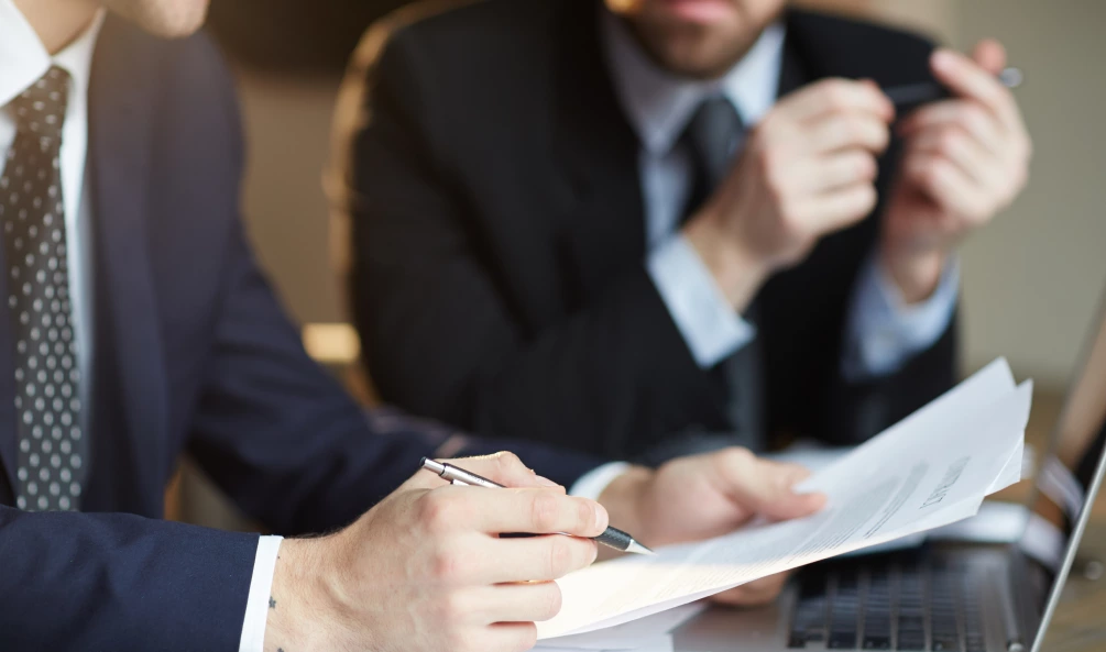 Two men in business suits are sitting at a desk reviewing documents. One is holding a piece of paper and a pen, while the other appears to be pointing or holding another item. A laptop is also on the desk.