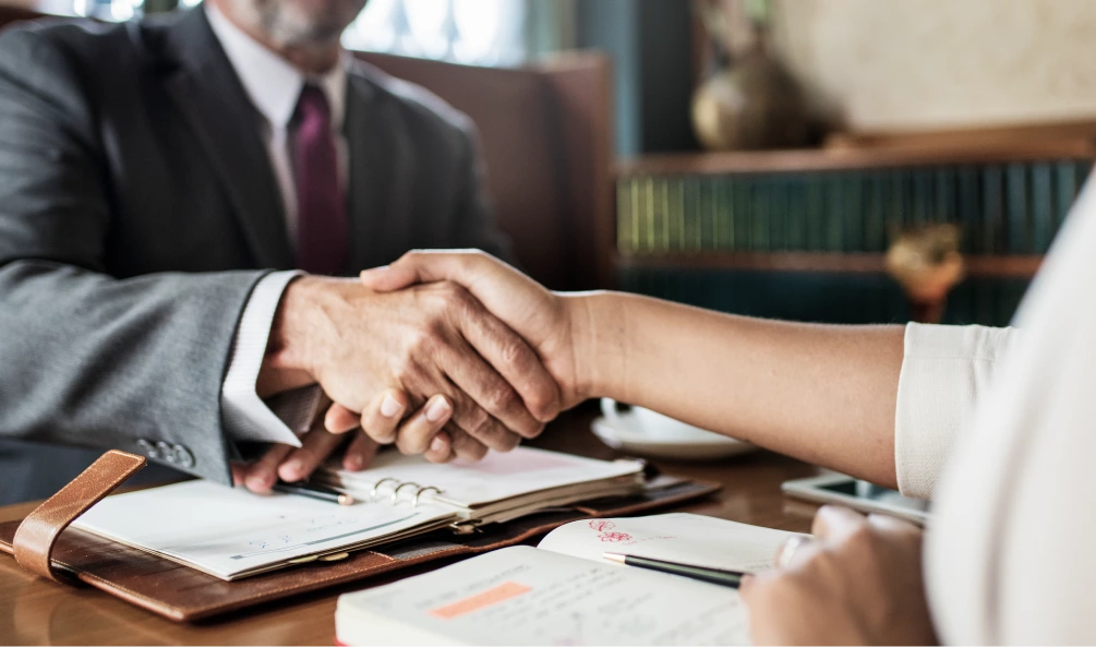 Two people, one in a suit, shake hands across a table with open notebooks and a planner, indicating a business meeting or agreement.