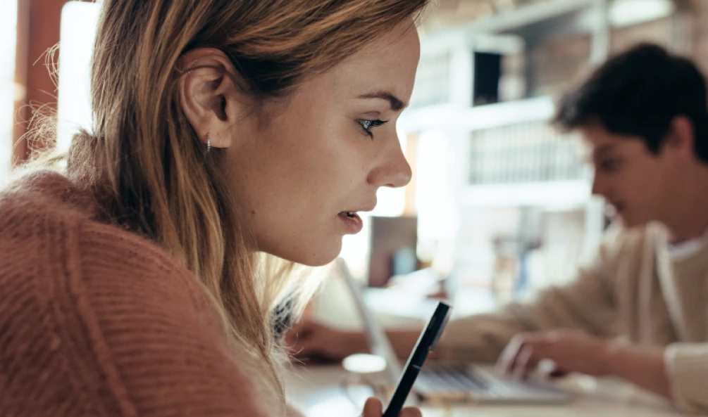 A woman focuses intently on a task while holding a pen, with a man using a laptop in the blurred background. They are in a well-lit indoor setting.