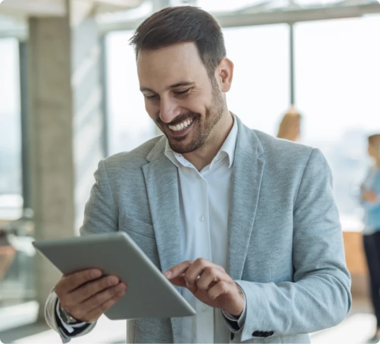 A man in a light gray blazer and white shirt smiles while using a tablet in a bright, modern office setting with large windows.