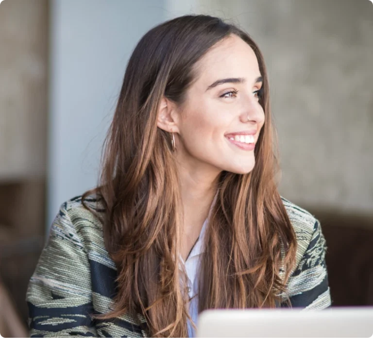 A woman with long brown hair smiles and looks to the side. She wears a patterned jacket and sits in front of a partially visible laptop.