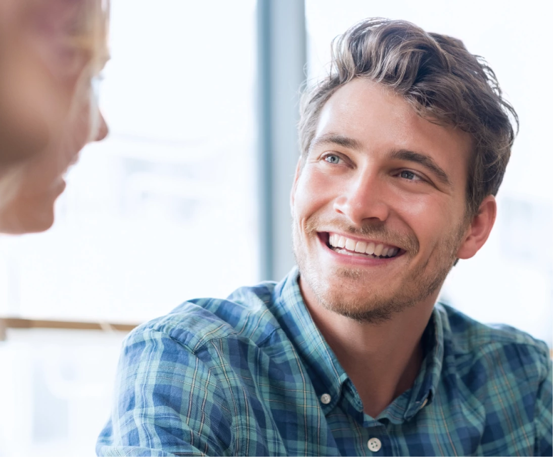 A man with wavy hair and a plaid shirt smiles warmly while looking at another person who is partially visible.