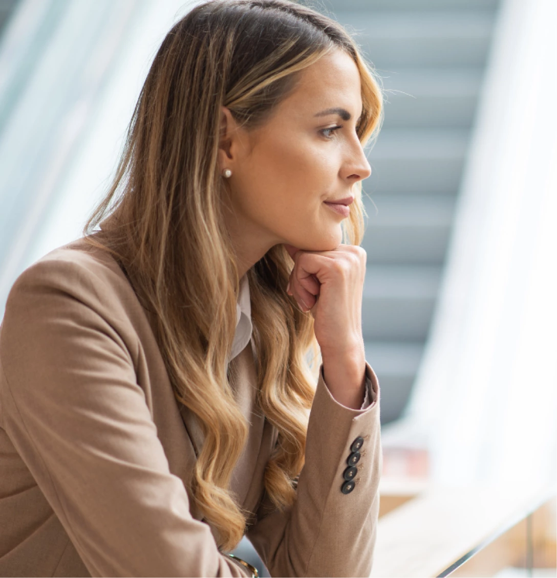 A woman with long blonde hair wearing a beige blazer is sitting indoors, resting her chin on her hand while looking to the side.