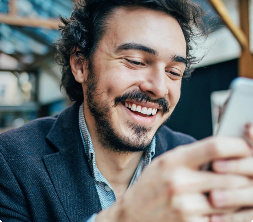 A person with curly hair and a beard is smiling while looking at their smartphone. They are wearing a jacket and a light-colored collared shirt.