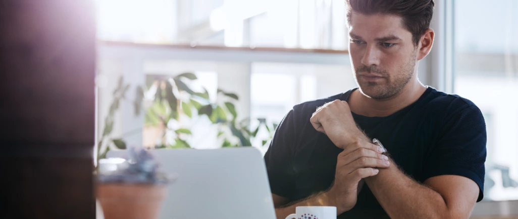 Man in a black shirt looking at his laptop screen with a serious expression, sitting at a desk with a coffee mug and a plant in the background.