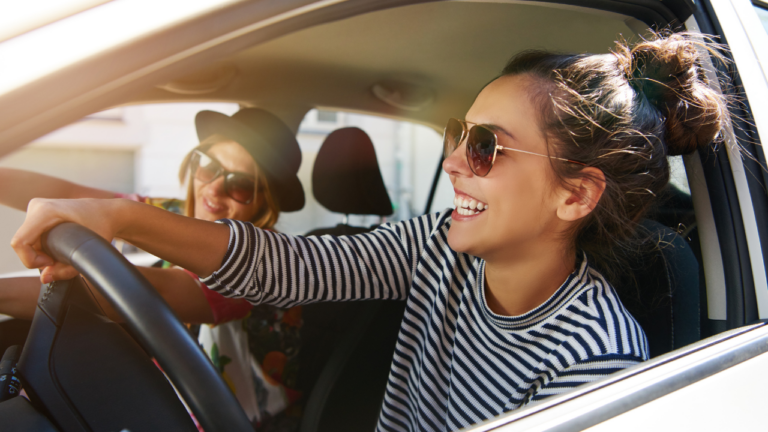 Two people are sitting in a car, smiling. The person in the driver's seat, sporting sunglasses and a striped shirt, exudes confidence possibly from recently mastering Texas Drivers Licenses: Everything You Need to Know. The passenger complements the vibe with a hat and sunglasses.