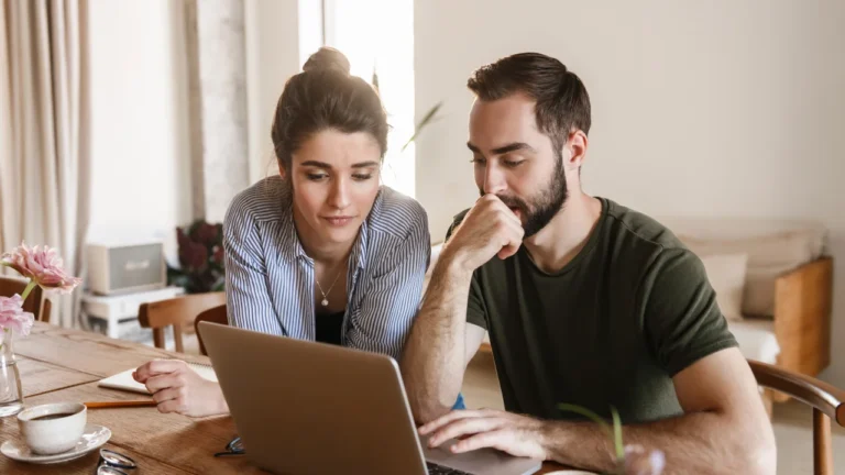 Two people sit at a wooden table, looking intently at a laptop. The woman wears a striped shirt, and the man wears a green shirt. The table has flowers, a mug, and papers. In their search for an Austin DWI lawyer, they seem deeply engrossed in finding the right legal representation.