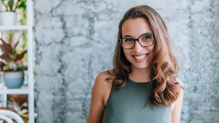 A smiling woman with long brown hair and glasses, wearing a sleeveless green top, stands in front of a textured gray wall. Potted plants are visible on a shelf in the background, subtly enhancing the warm atmosphere often seen in an Austin DWI lawyer's office.