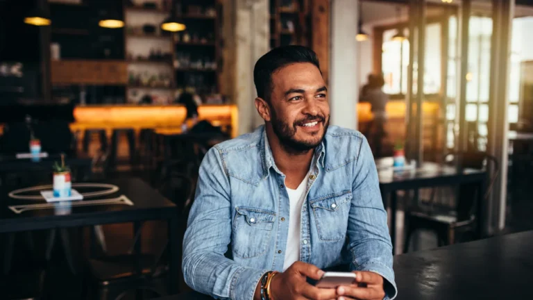 A man in a denim shirt sits at a table in a café, holding a smartphone and smiling, as he reads the latest post from his favorite San Antonio DWI lawyer.