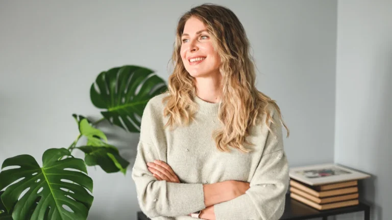 A person with long, wavy hair stands indoors, smiling with arms crossed. They are wearing a light gray sweater. There are green plants and stacked books in the background, making it an inviting space—perhaps the home office of an San Antonio DWI lawyer.