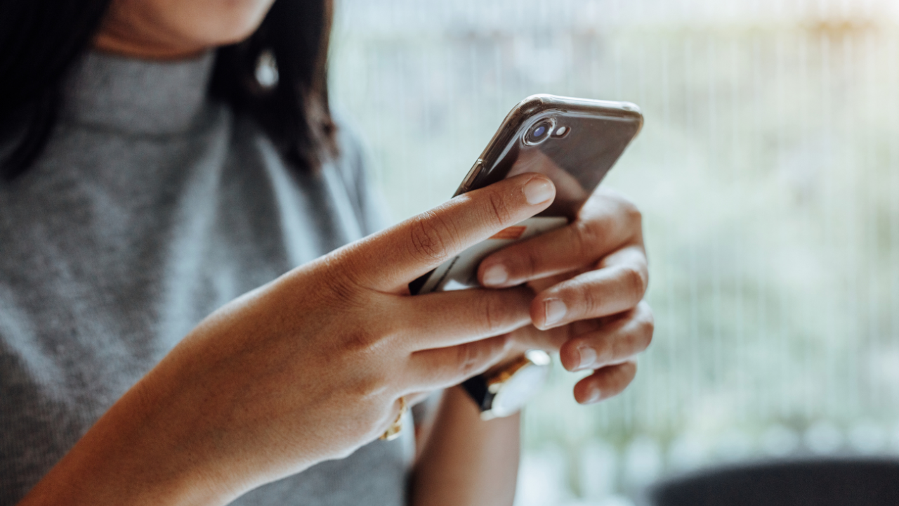 Person holding a smartphone and tapping the screen with both hands, wearing a gray top, with a blurred background, perhaps searching for information on topics like "Can You Get Early Release From Probation In Texas?