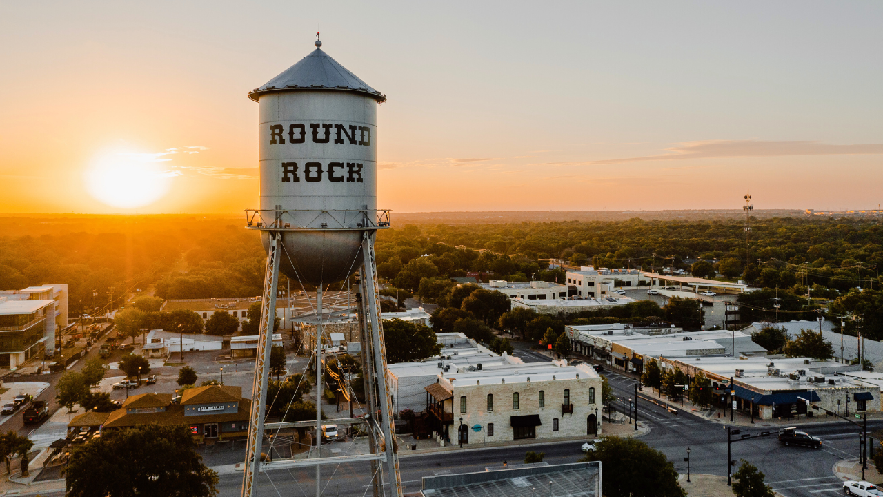 Aerial view of a water tower labeled "Round Rock" with a town and trees below, set against a sunset sky. In the heart of this picturesque scene, you'll also find trusted services including Round Rock DWI Lawyer professionals ready to assist the community.