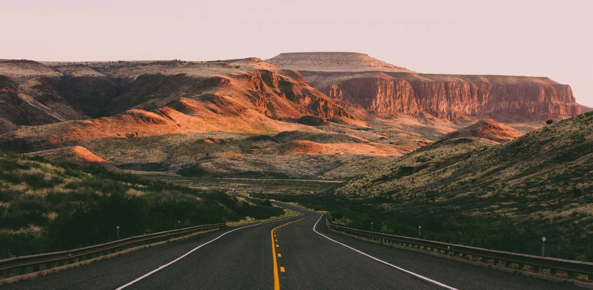 A paved road leads through a desert landscape with rocky hills and plateaus under a light pink sky, reminiscent of the vast stretches one might drive to visit a Corpus Christi DWI lawyer.