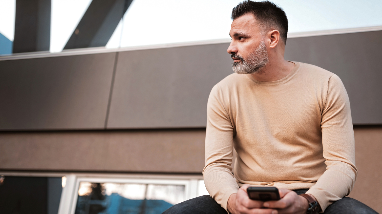 A man with a beard sits outside, holding a smartphone and looking to the left. He is dressed in a beige sweater and dark pants. Behind him, the modern building structure adds an urban vibe as he casually ponders questions like how much does insurance go up after DUI in Texas.