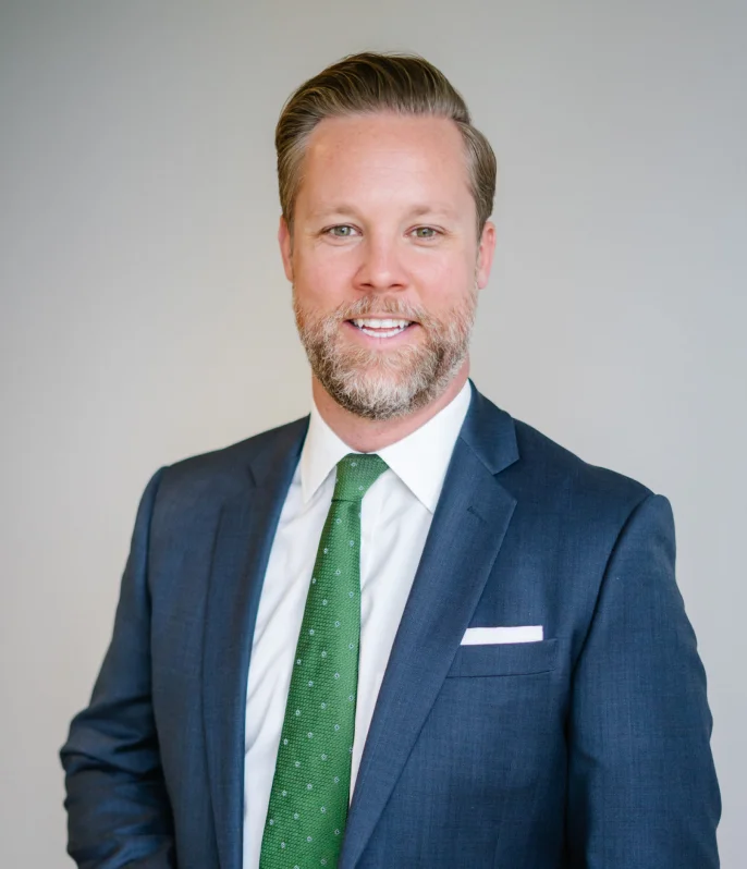 A man with a beard and mustache is smiling, wearing a dark blue suit, white shirt, and green tie, standing in front of a plain light-colored background. About Trey Porter.