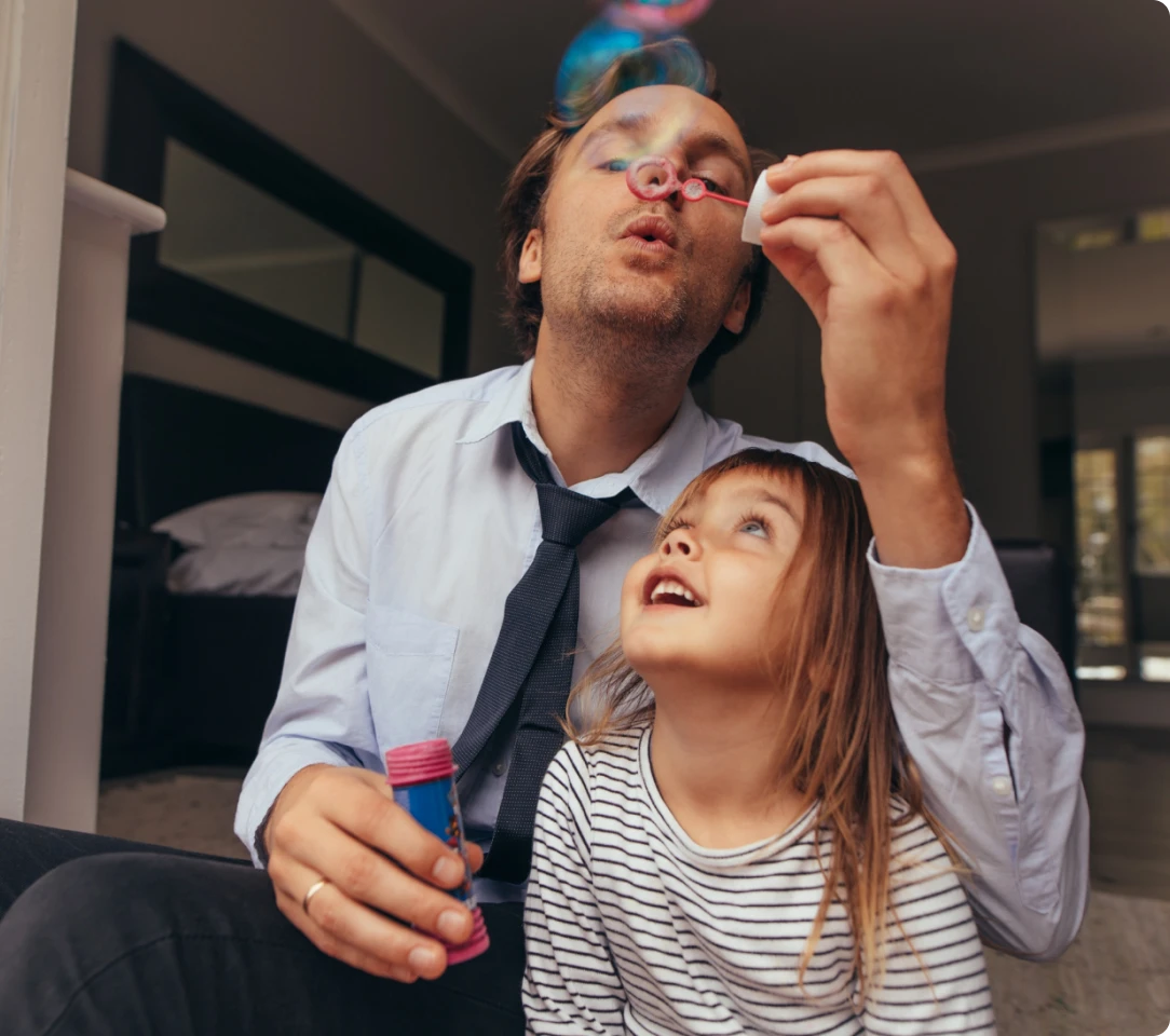 An adult and a child are sitting indoors. The adult is blowing bubbles while the child, wearing a striped shirt, looks up at the bubbles in anticipation, creating a moment of simple joy that contrasts sharply with the serious tones of Texas DWI Attorney Reviews.