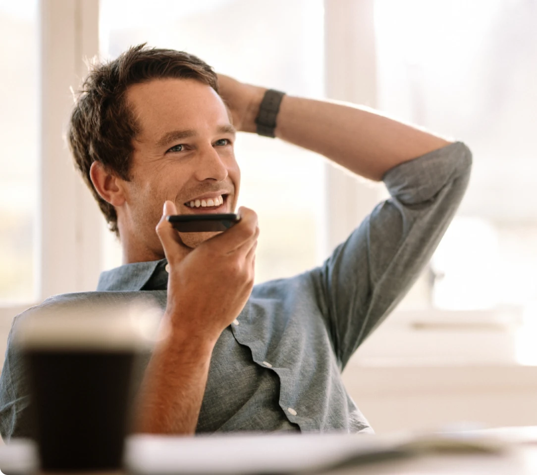 A smiling man with dark hair and a blue shirt holds a smartphone to his mouth while sitting back with one hand behind his head, perhaps reading Texas DWI Attorney Reviews.