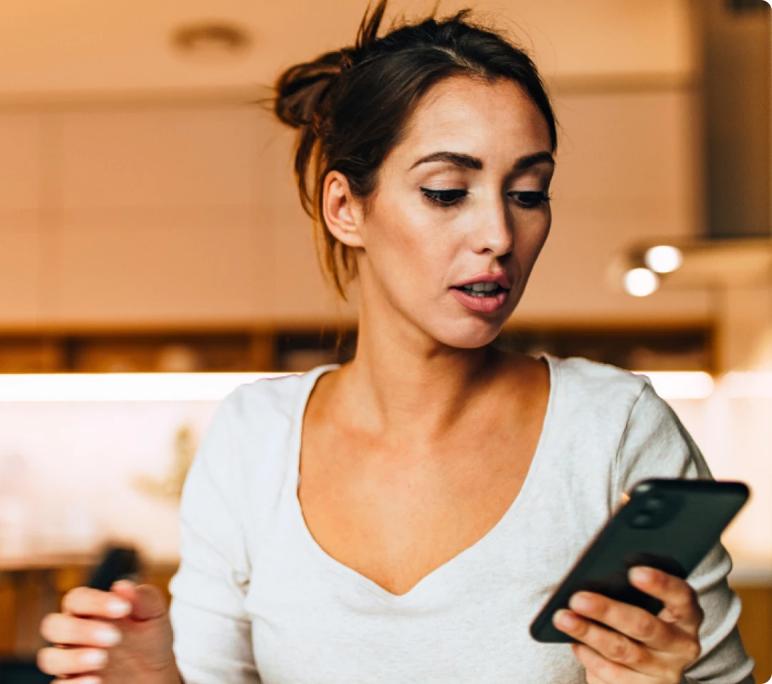 A woman in a white top, with her hair tied up, looks at her smartphone in a kitchen setting, possibly reading Texas DWI Attorney Reviews.