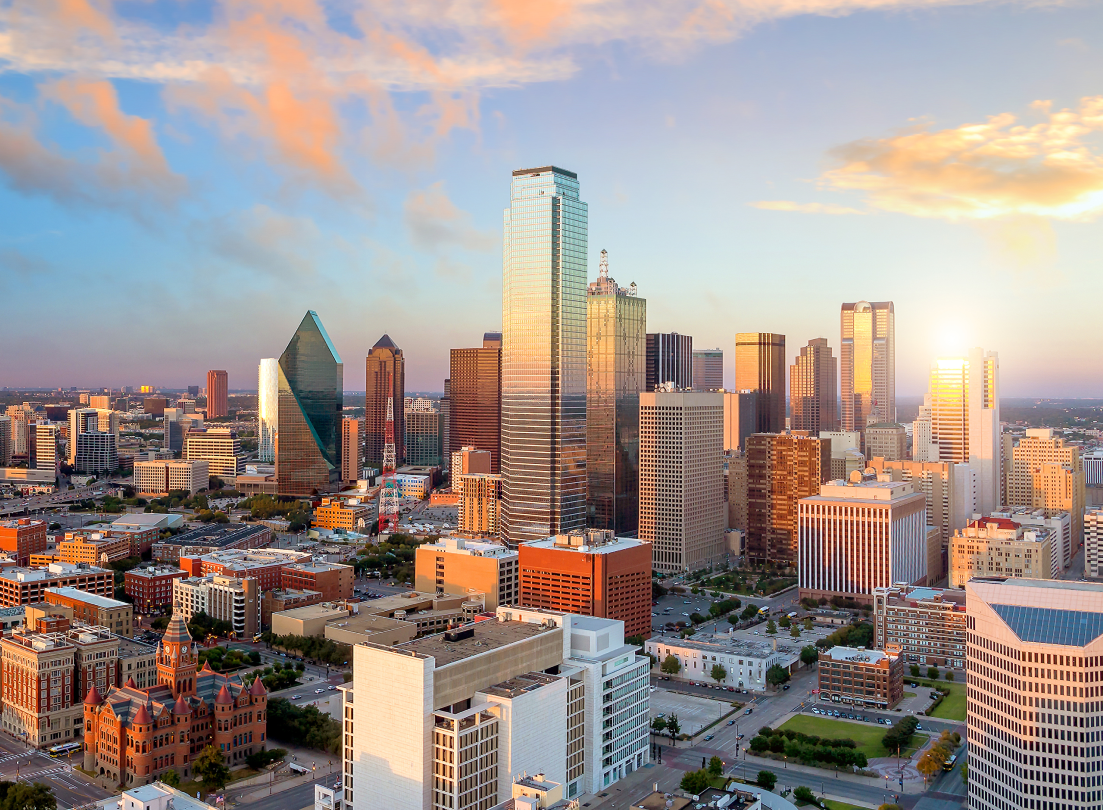 Aerial view of a city skyline at sunset, featuring a mix of modern skyscrapers and historic buildings under a partly cloudy sky.