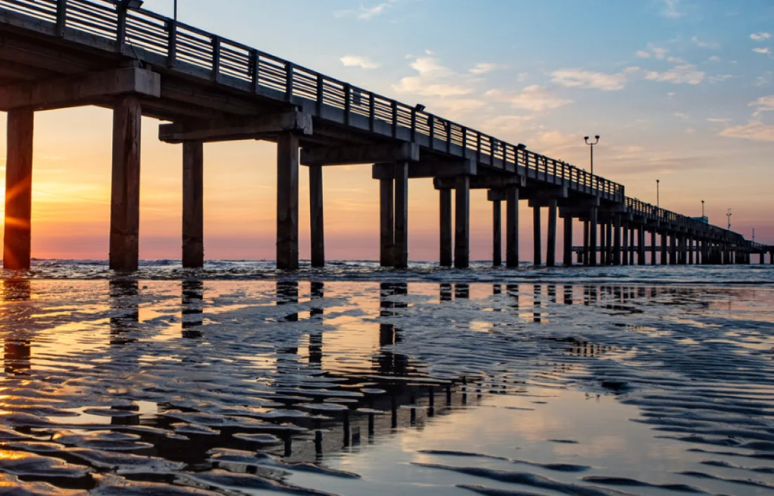 A long pier extends over the ocean at sunset. The sky is colorful with orange, pink, and blue hues. The wet sand in the foreground reflects the pier and the sky.