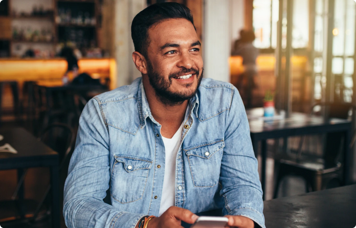 A man in a denim jacket is sitting at a table in a cafe, smiling while holding a smartphone as he browses Texas DWI Attorney Reviews.