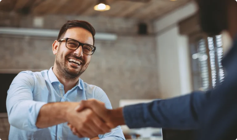 Two people shaking hands in an indoor office environment. One person, smiling, wears glasses and a light blue shirt.