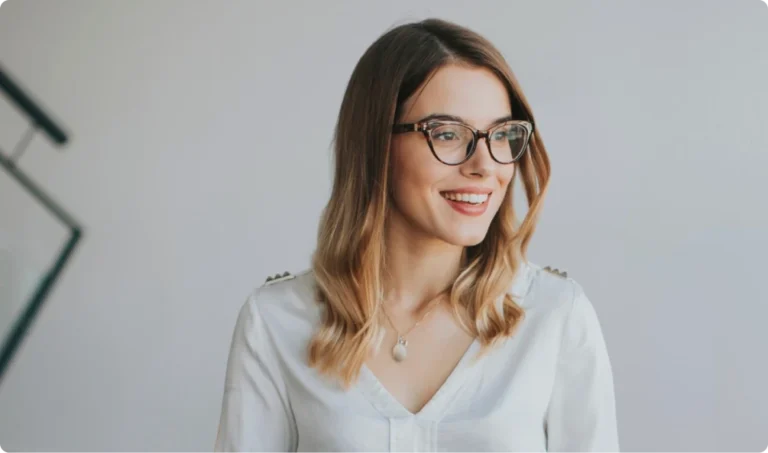 A woman with shoulder-length blonde hair and glasses smiles while looking to the side, wearing a white blouse with a visible pendant necklace.