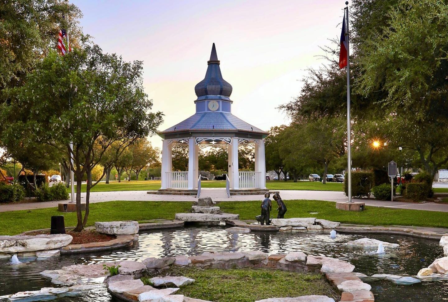 A small blue and white gazebo stands in the center of a park, surrounded by trees and flags, with a circular pond featuring a stone pathway in the foreground—a serene spot, contrasting sharply with the bustling nearby offices of Blanco DWI Lawyer.