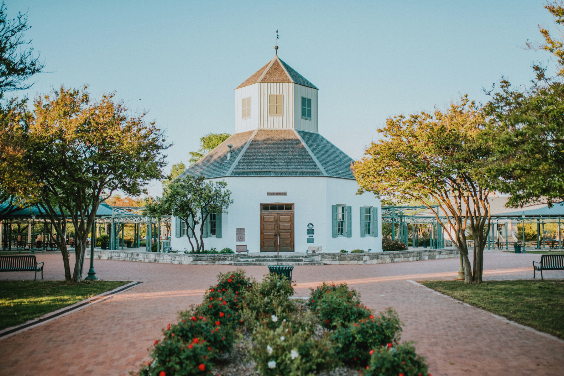 A small, white, octagonal building with a central entrance is surrounded by walkways, gardens, and trees under a clear blue sky, much like the inviting office of a dedicated Fredericksburg DWI Lawyer.