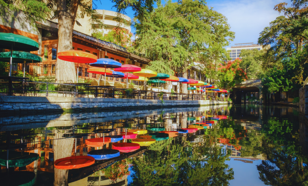 Colorful umbrellas line a riverside dining area on a sunny day, reflecting in the calm water below. Trees and buildings surround the area, creating a picturesque scene. This charming locale is just one of the many beautiful spots you can enjoy in San Antonio DWI Lawyer neighborhood.