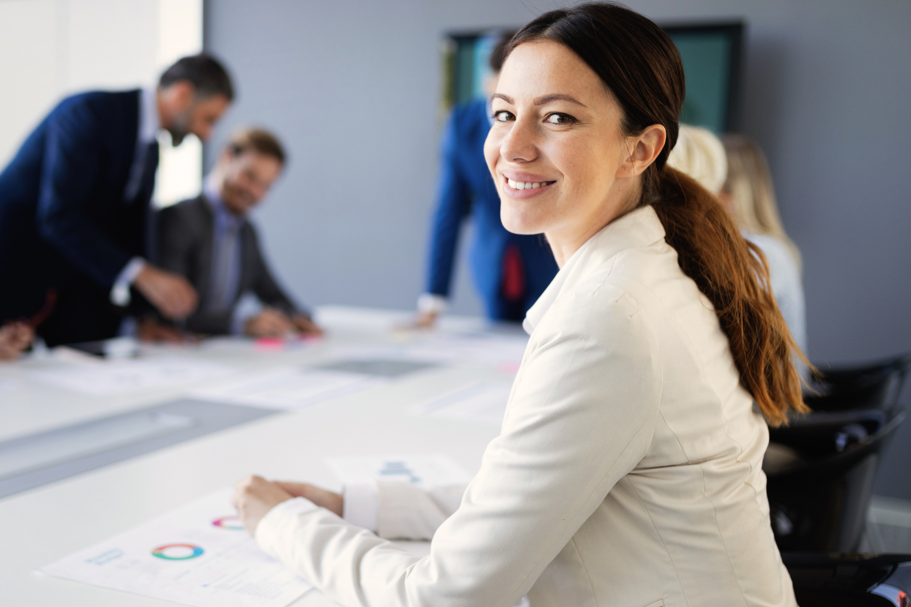 A woman in a white blazer sits at a conference table, smiling at the camera. In the background, people are engaged in a discussion about crimes expunged in Texas, with documents spread out on the table.