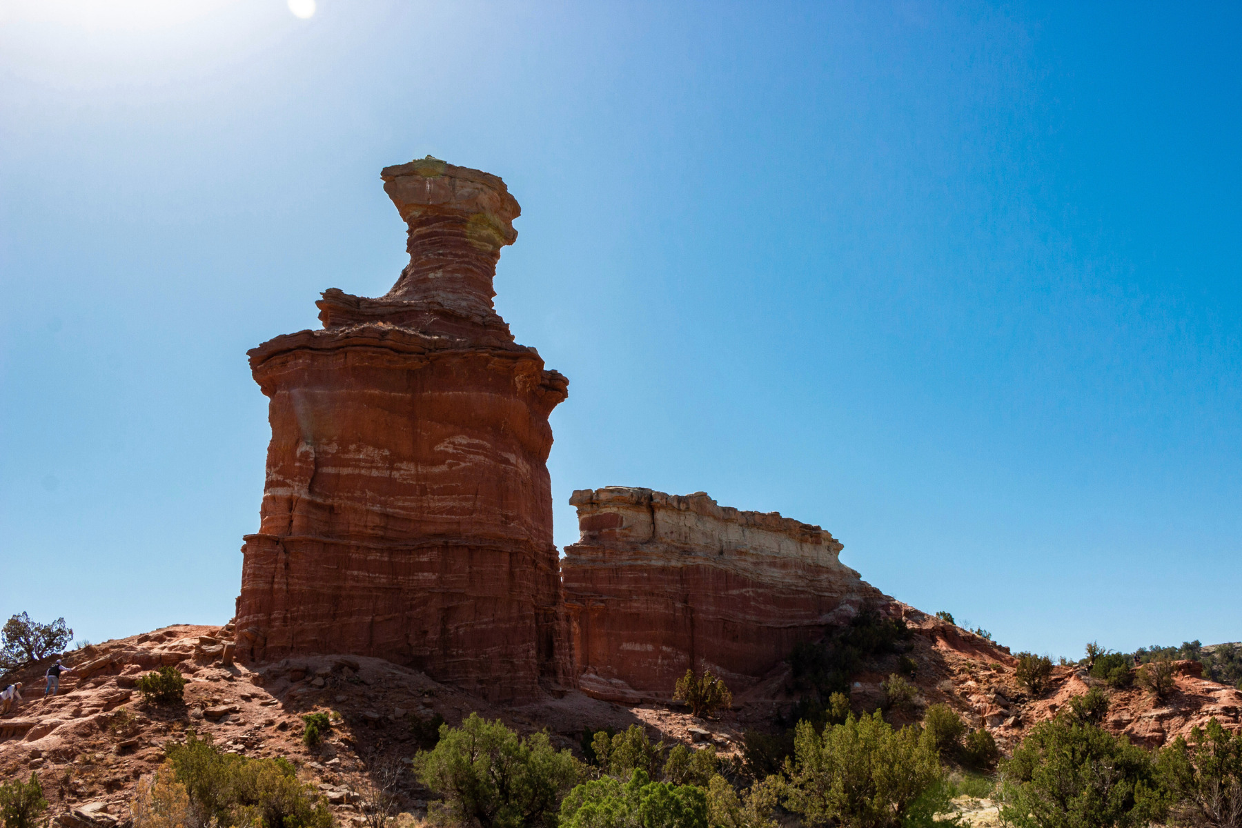 A tall, layered red rock formation stands under a clear blue sky, akin to the steadfast reputation of an Amarillo expungement lawyer, surrounded by shrubs and smaller rock formations.