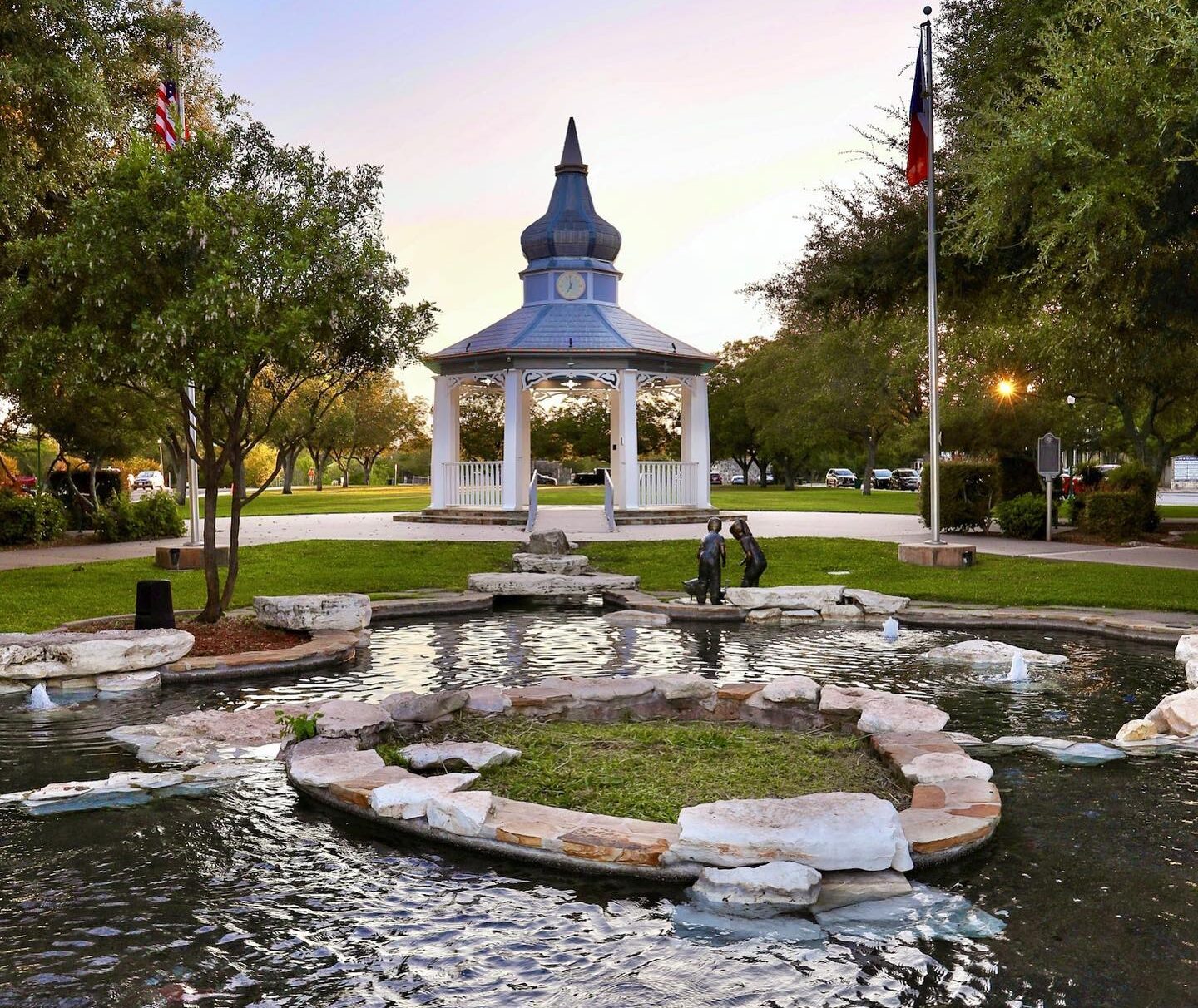 A park scene with a pond and a small stone island in the foreground evokes tranquility. A gazebo with a clock tower rises in the background, surrounded by trees, while two flags flutter above. It’s hard to imagine such serenity disturbed by any occurrence of public intoxication in Boerne.