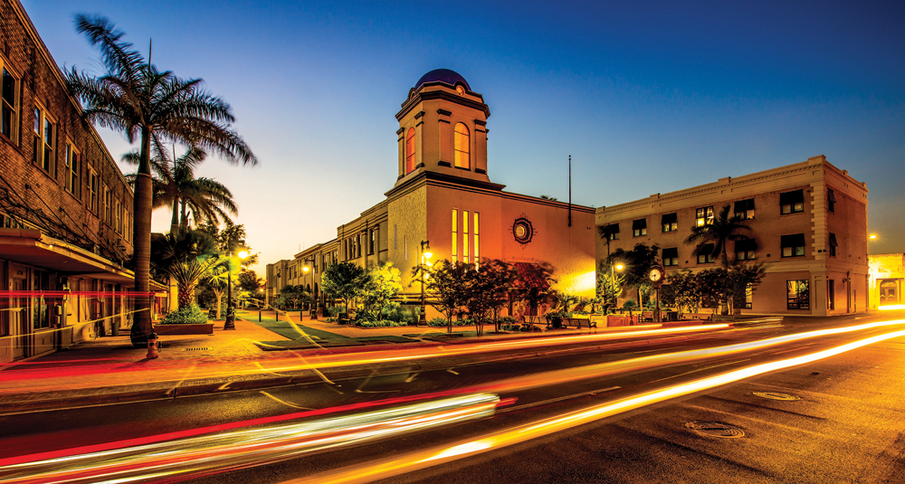 As twilight descends, light trails from moving vehicles dance across the street scene. A historic building with a towering presence is surrounded by palm trees and adjacent structures, much like the distinguished work of a Brownsville Expungement Lawyer navigating through legal pathways.