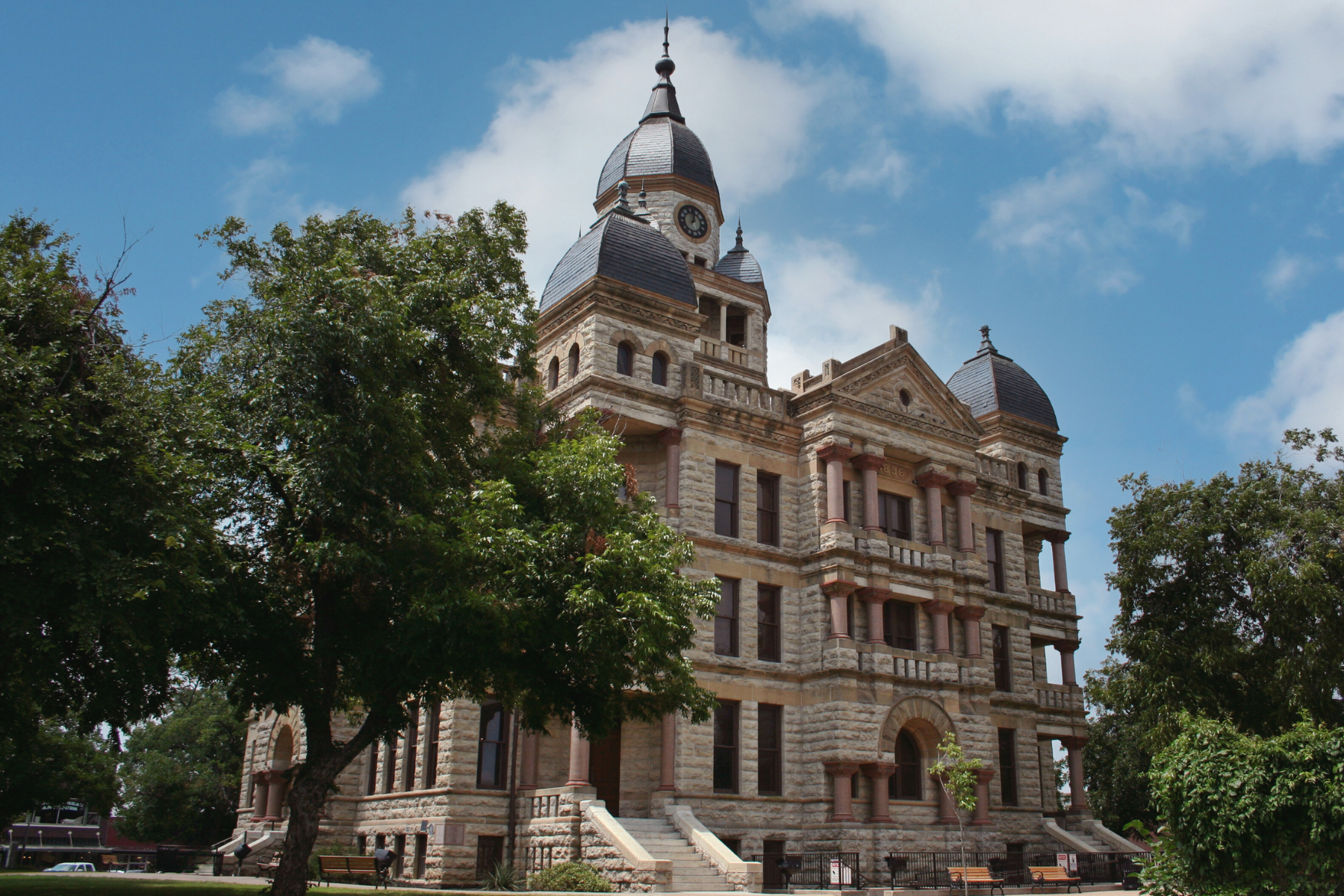 The historic stone courthouse, with its majestic clock tower and tree-lined entrance, stands proudly under a partly cloudy sky, reminiscent of days when public intoxication in Denton was a lively debate.