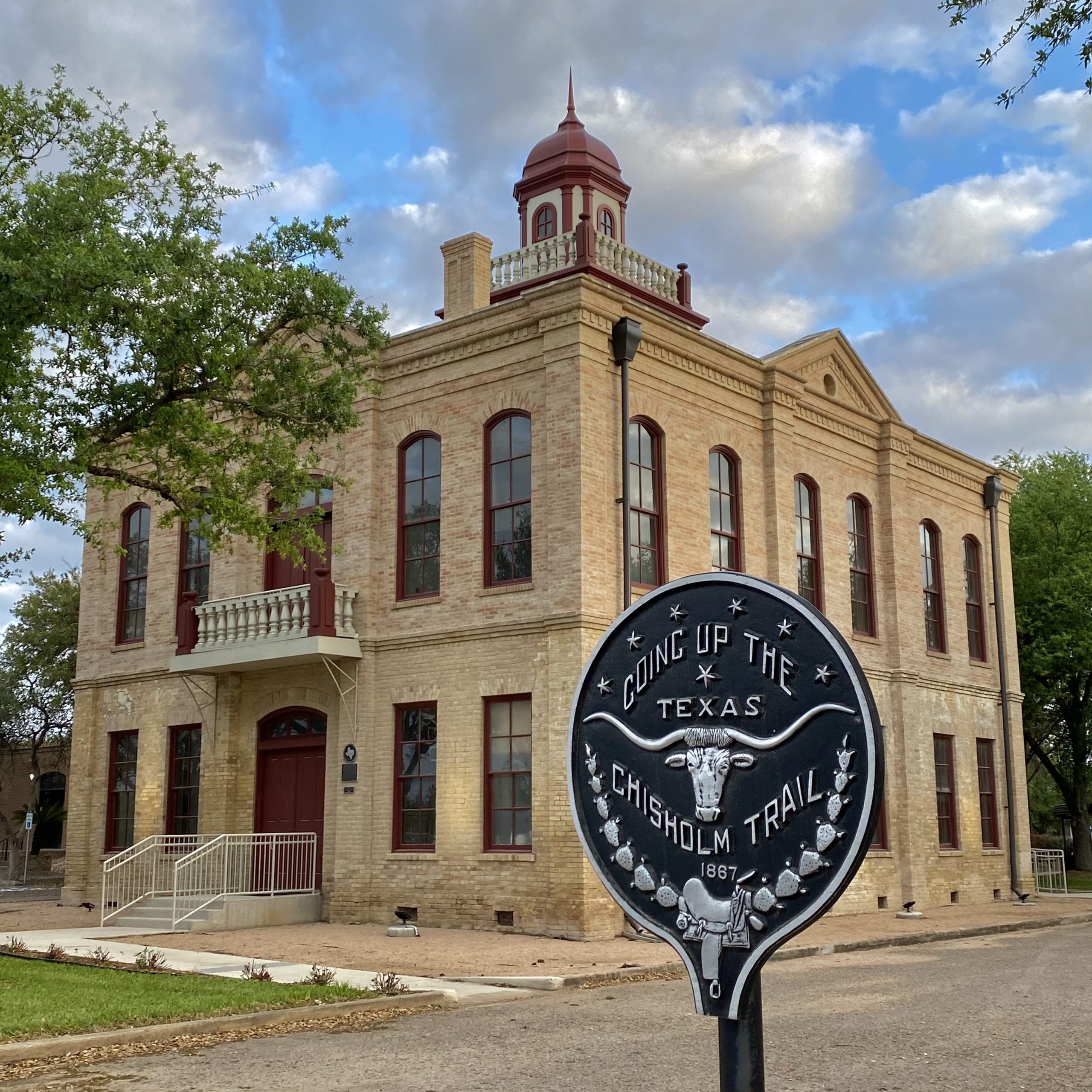 Historic brick building with a central tower and red accents under a cloudy sky. In front, an Edinburg Expungement Lawyer's office stands near a Chisholm Trail marker dated 1887.