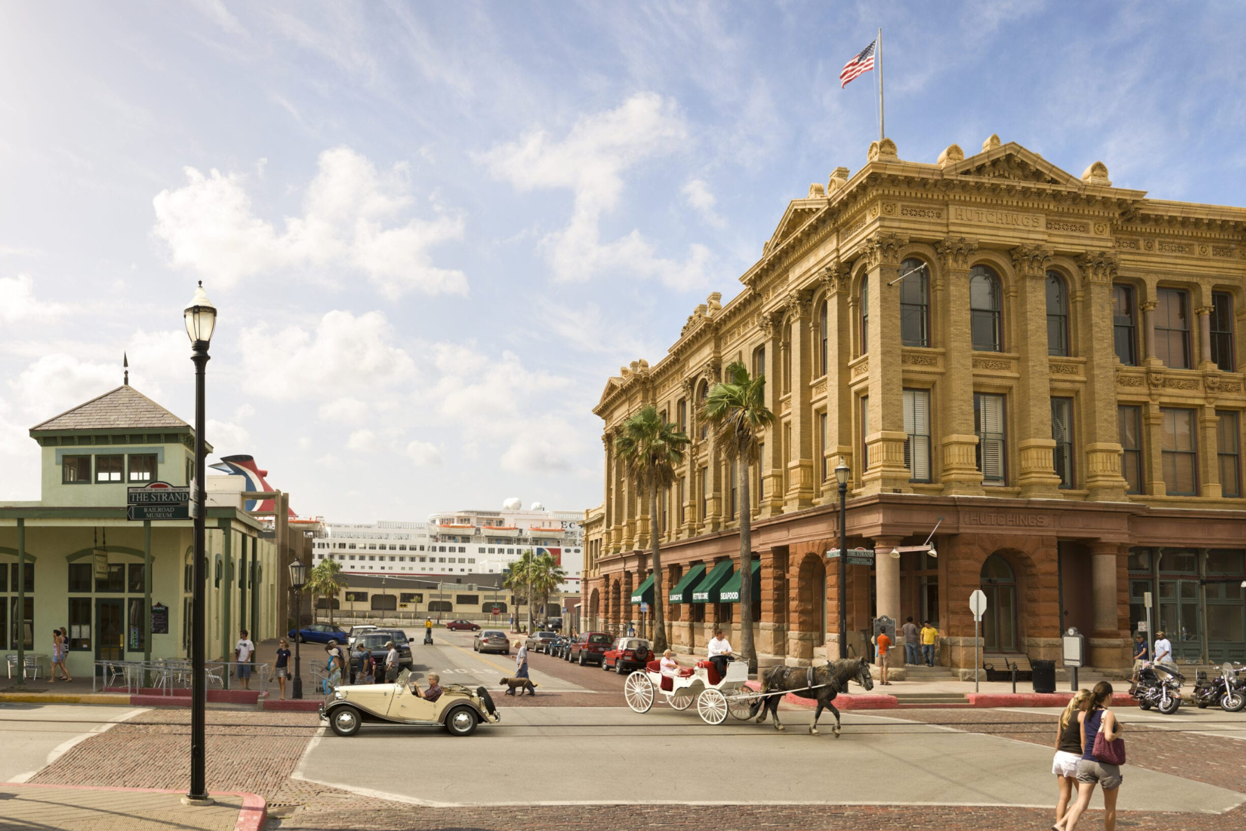 In a historic street scene, a horse-drawn carriage and vintage car meander past pedestrians near an ornate brick building with a U.S. flag. This picturesque moment stands in stark contrast to tales of public intoxication in Galveston's equally lively streets.