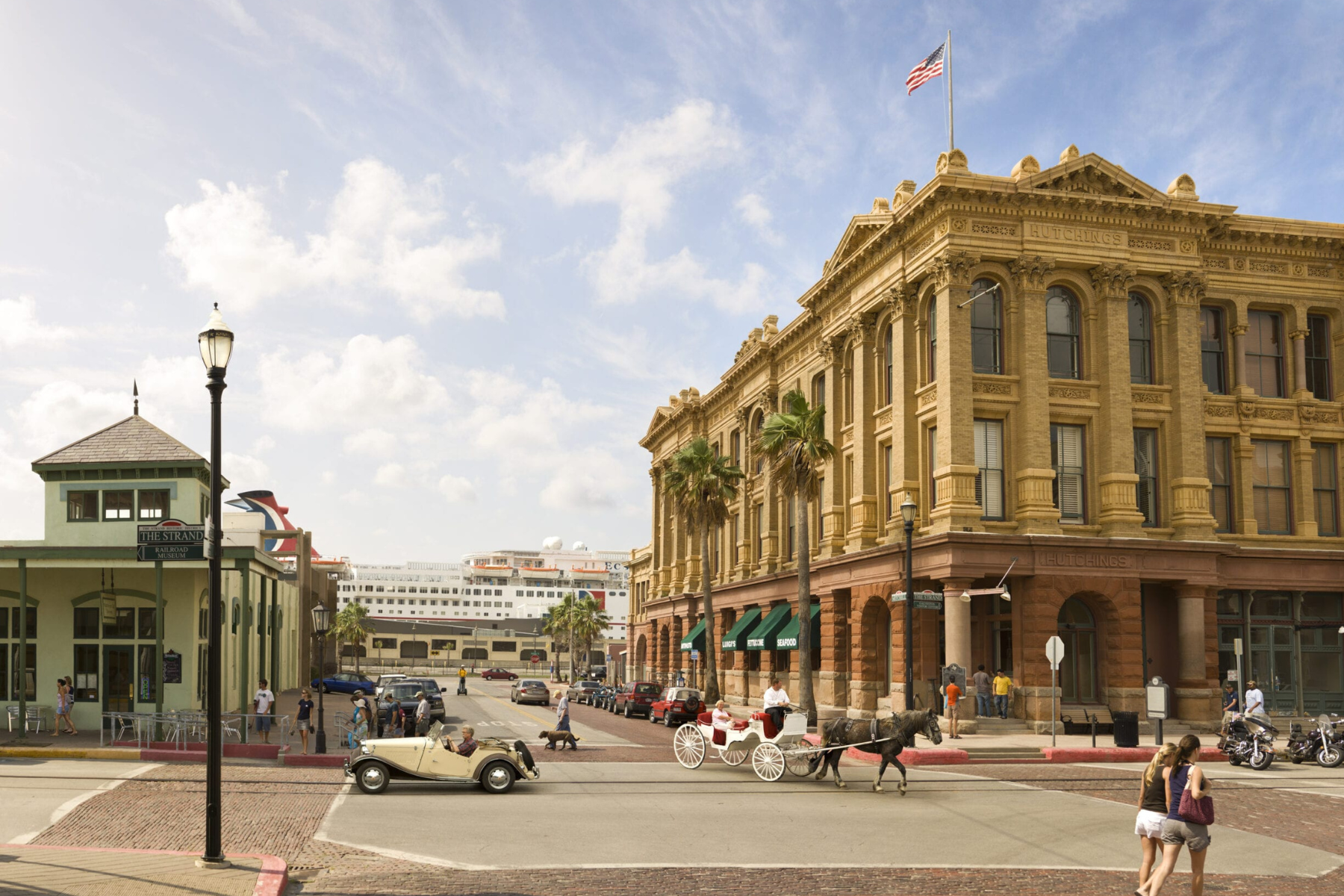 Historic street scene with a vintage car and horse-drawn carriage in front of a large, ornate building, an American flag waving proudly above. This picturesque setting evokes a sense of timeless charm, much like the reliable service you'd expect from a Galveston expungement lawyer.