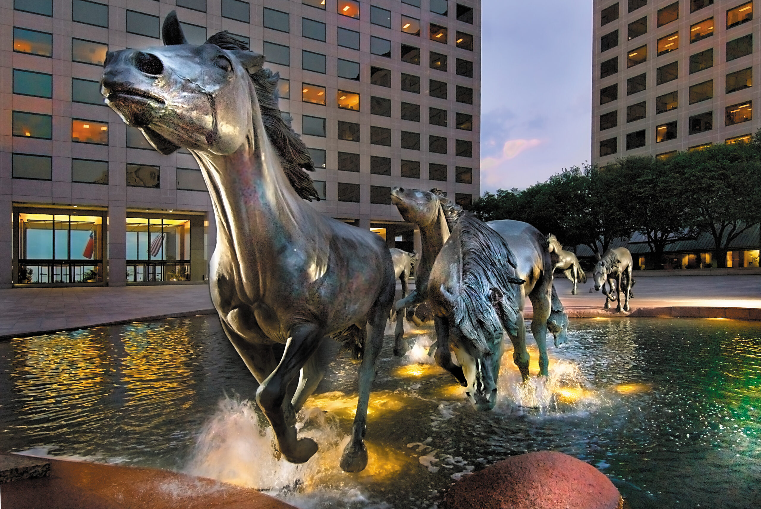 A statue of wild horses, akin to the momentum one might feel with a skilled Irving Expungement Lawyer, gallops through water between two tall buildings at dusk.