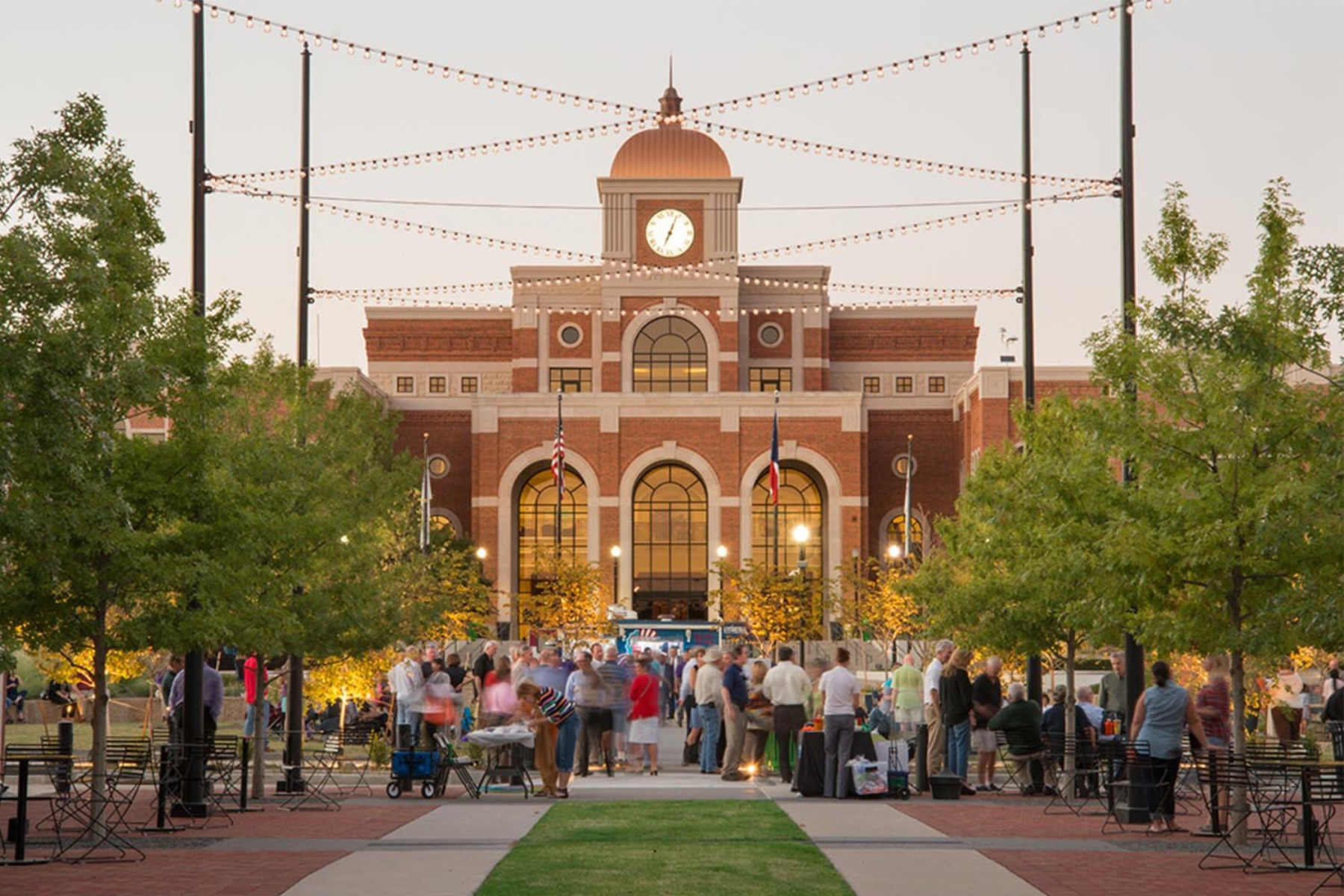 A large building with a dome and clock tower looms in the background, while a lively crowd gathers in front, surrounded by trees and string lights. It's almost as if they're celebrating, perhaps unaware of the public intoxication issues that have been a concern in Lewisville.