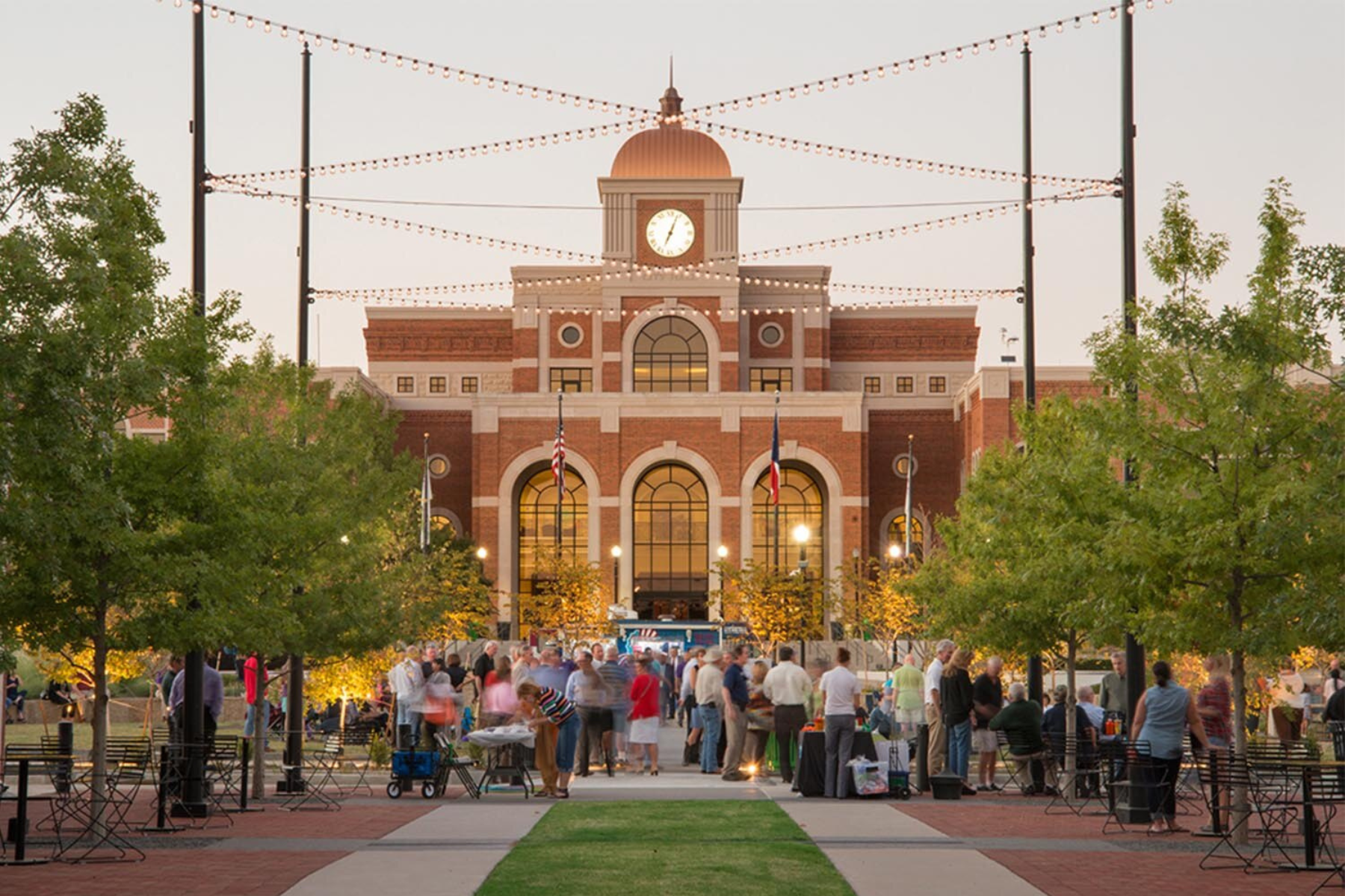 A crowd gathers in front of a large red-brick building, resembling the prestigious office of a Lewisville Expungement Lawyer, with a central clock tower. It's surrounded by trees and decorated with string lights.