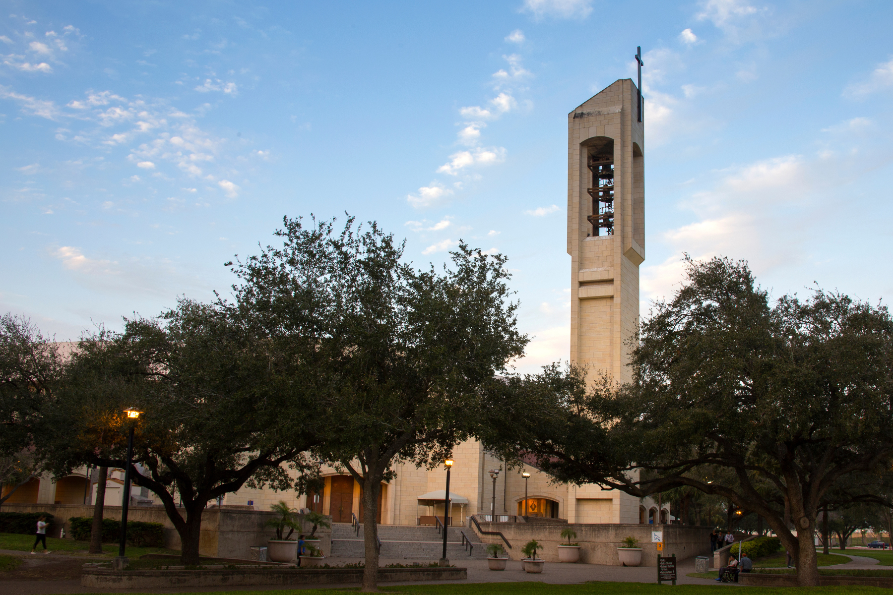 A tall bell tower stands against a blue sky with scattered clouds, surrounded by trees and a walkway where a few people stroll leisurely, offering no hint of the public intoxication in McAllen.