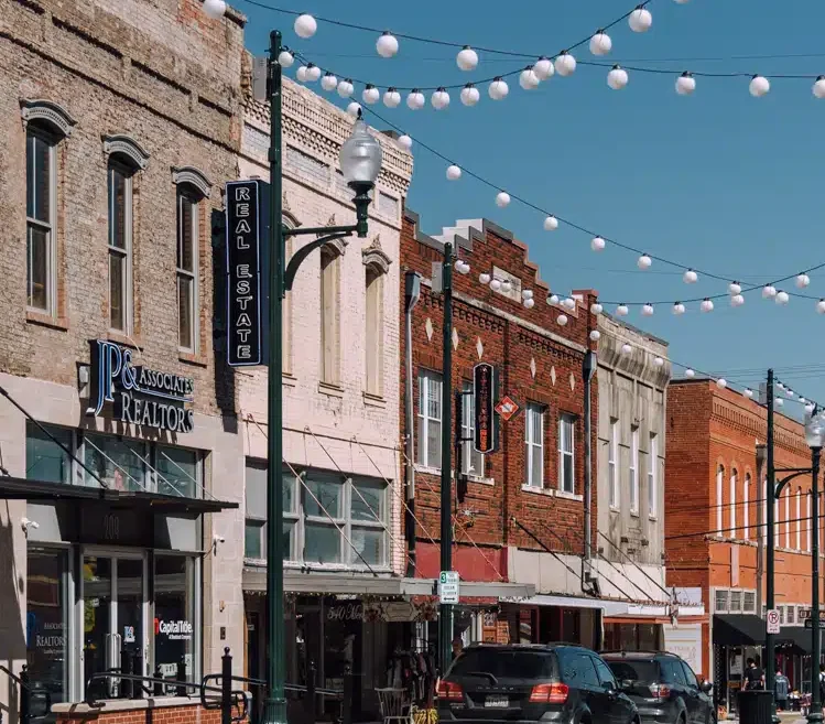 A street view of a small town with brick buildings, featuring a realtor's office and a café. Cars are parked along the road under string lights, capturing an unexpected charm that's worlds away from the hustle found in places like McKinney.