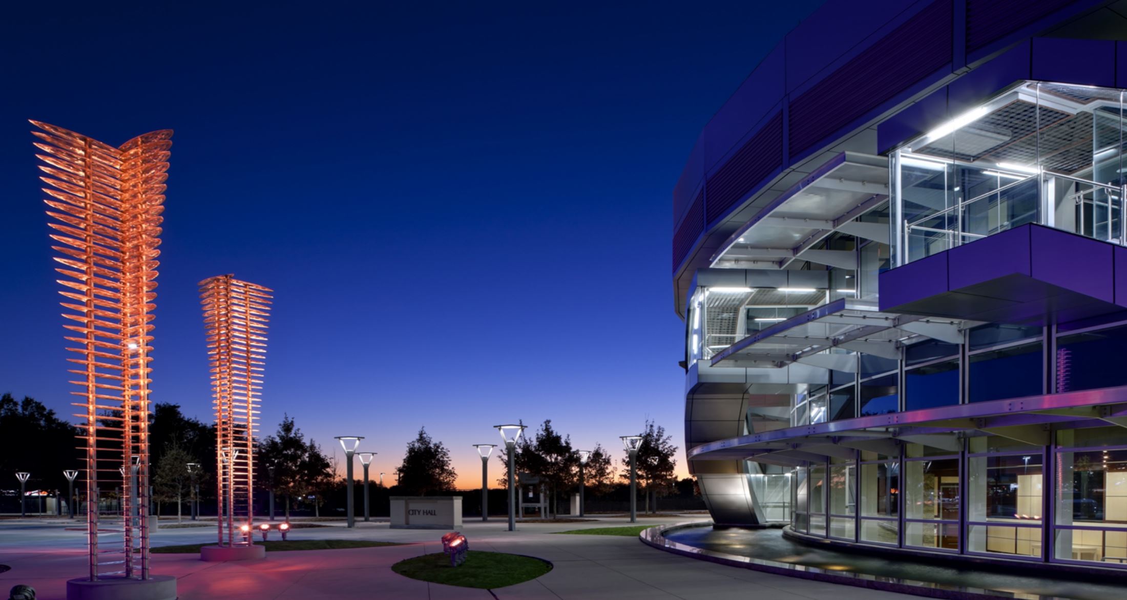 As dusk falls over the modern building in Mesquite, its glass facade shimmers against a twilight sky while illuminated art sculptures add a mesmerizing touch.